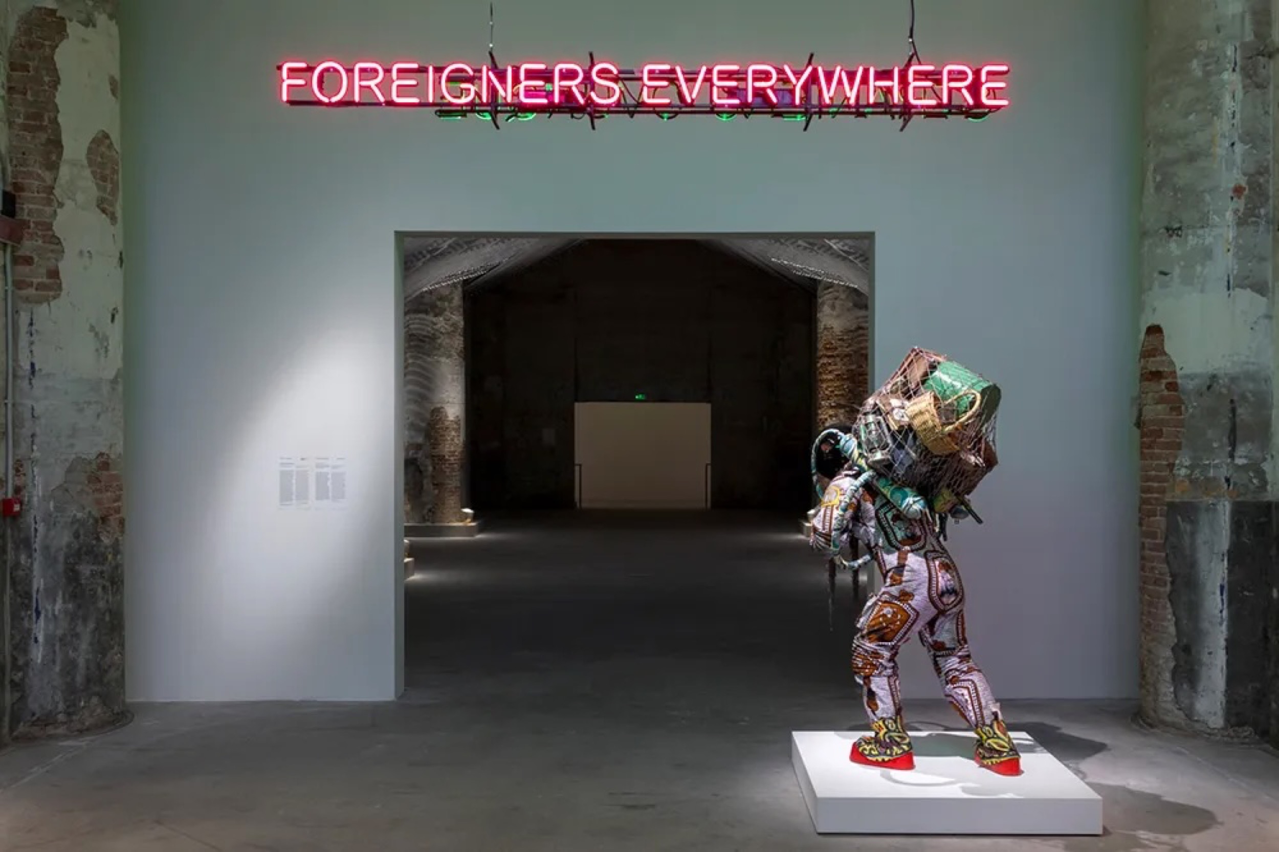<p>Installation view of <em>Foreigners Everywhere</em> (2004–) by Claire Fontaine and <em>Refugee Astronaut VIII</em> (2024) by Yinka Shonibare at the 2024 Venice Biennale. Photography by Marco Zorzanello. Image courtesy of the artist and La Biennale di Venezia.</p>